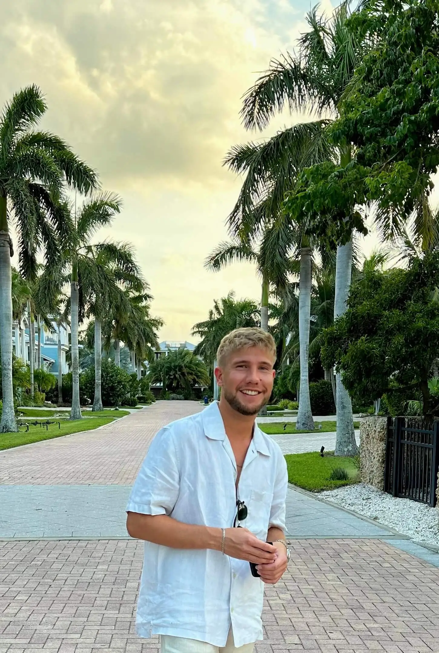Smiling man in white shirt standing on a palm-lined brick pathway.